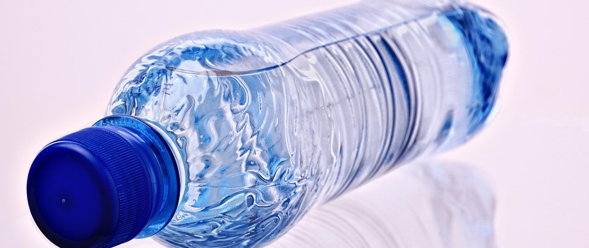 Woda w plastikowej błękitnej butelce z niebieską plastikową nakrętką. Woda w butelce znajduje się w pozycji leżącej. Białe tło. Widoczne lustrzane odbicie leżącej butelki.