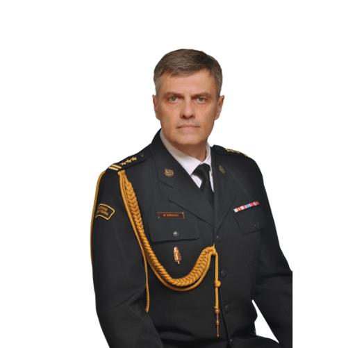 Portretowy, studyjny wizerunek mężczyzny w średnim wieku w ciemnym, galowym mundurze służbowym. Mężczyzna ma krótkie, siwiejące włosy i poważny wyraz twarzy. Na mundurze widoczne są odznaczenia, złote sznury ozdobne na ramieniu oraz pagony z oznaczeniami stopnia. Na piersi znajduje się imiennik z nazwiskiem. Na rękawie munduru widoczna jest naszywka z napisem w języku polskim. Tło zdjęcia jest jasne i neutralne, utrzymane w odcieniach szaro-niebieskich.