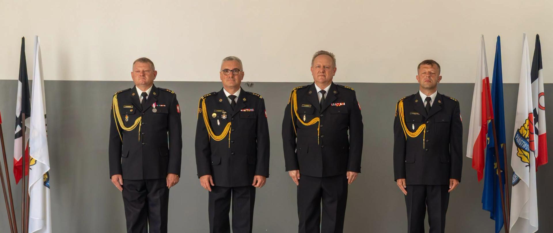 Oficjalne zdjęcie przedstawiające czterech funkcjonariuszy Państwowej Straży Pożarnej stojących w baczności w jednym rzędzie. Mężczyźni są ubrani w ciemnogranatowe mundury wyjściowe z białymi koszulami, czarnymi krawatami oraz ozdobnymi, złotymi sznurami galowymi (akselbantami) przypiętymi do prawego ramienia. Na ich piersiach widoczne są baretki odznaczeń oraz medale. Strażacy stoją w pomieszczeniu na tle szaro-białej ściany. Po lewej i prawej stronie kadru, za mężczyznami, ustawione są stojaki z flagami, wśród których widoczna jest biało-czerwona flaga Polski, flaga Unii Europejskiej oraz flagi z emblematami formacji strażackiej.