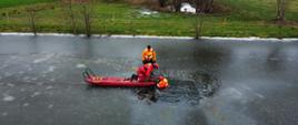Strażaka znajdującego się na saniach lodowych, podejmującego próbę ewakuacji drugiego strażaka znajdującego się w wodze w przeręblu. Za nimi stoi kierownik ćwiczeń sprawdzający poprawność wykonanej techniki.