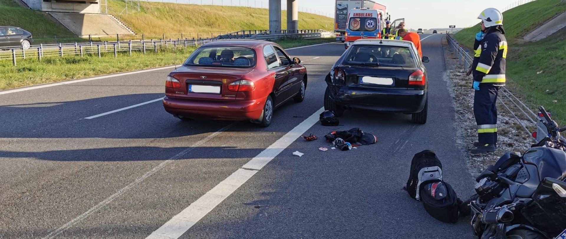 Zdjęcie obrazuje dwa samochody osobowe znajdujące się na jednym pasie ruchu drogi ekspresowej S7. Jeden z nich ma uszkodzony tylni zderzak. Na pasie awaryjnym o barierę energochłonną oparty jest uszkodzony motocykl. Na drugim planie widać karetkę pogotowia.