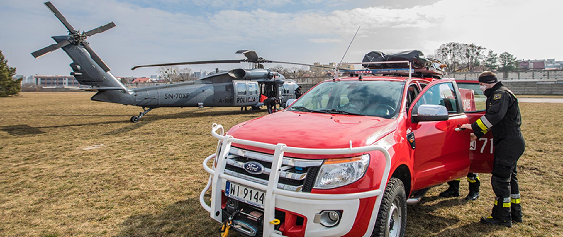 Wspólne ćwiczenia Policji i Państwowej Straży Pożarnej. Na pierwszym planie terenowy samochód pożarniczy. W tle biorący udział w ćwiczeniu śmigłowiec.