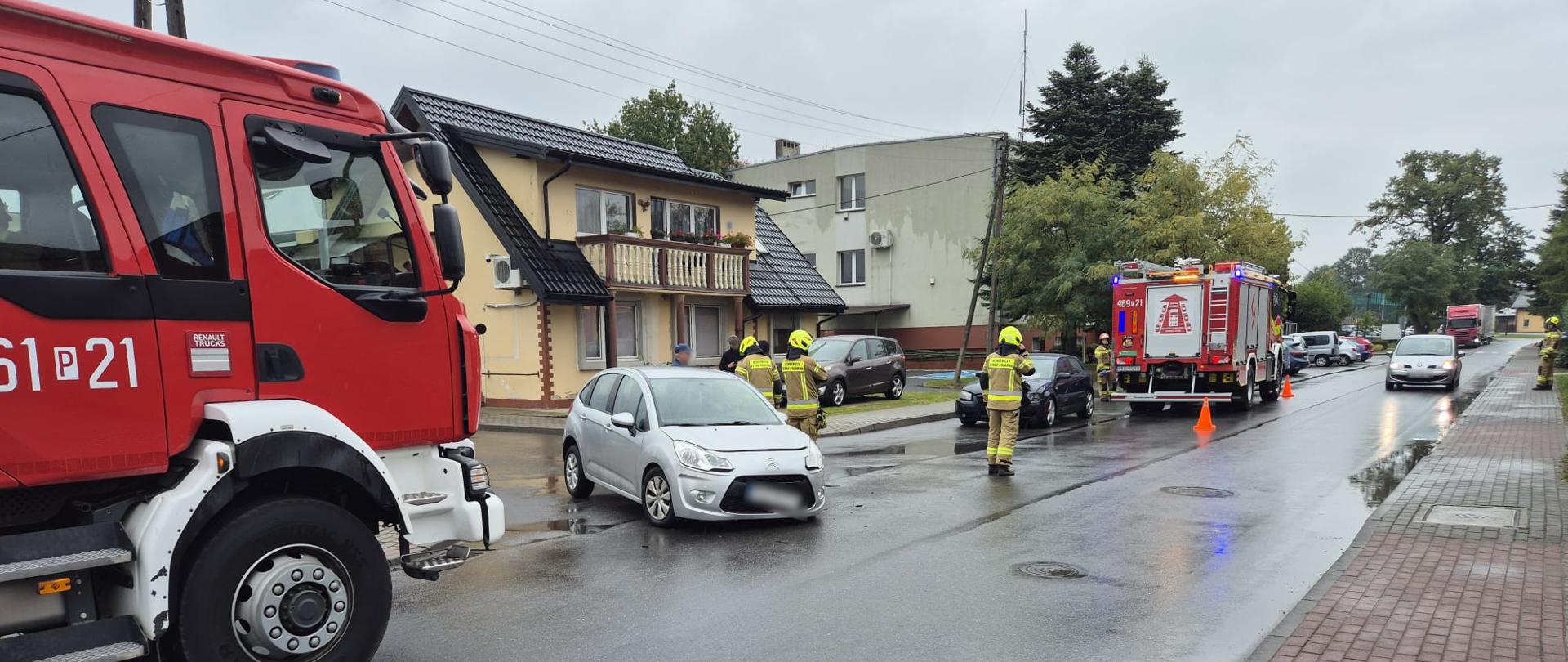 Na zdjęciu widać miejsce zdarzenia drogowego na mokrej, asfaltowej ulicy w mieście. Na pierwszym planie stoi czerwony wóz strażacki, a w dalszej części drogi znajduje się drugi pojazd straży pożarnej z włączonymi światłami ostrzegawczymi i ustawionymi pomarańczowymi pachołkami. Pomiędzy nimi stoi srebrny samochód osobowy z widocznymi uszkodzeniami z przodu. Na miejscu obecni są strażacy w żółtych strojach ochronnych i kaskach, którzy zabezpieczają teren. W tle widać budynki mieszkalne, zaparkowane samochody oraz chodnik z kostki brukowej. Pogoda jest deszczowa, a nawierzchnia drogi mokra.