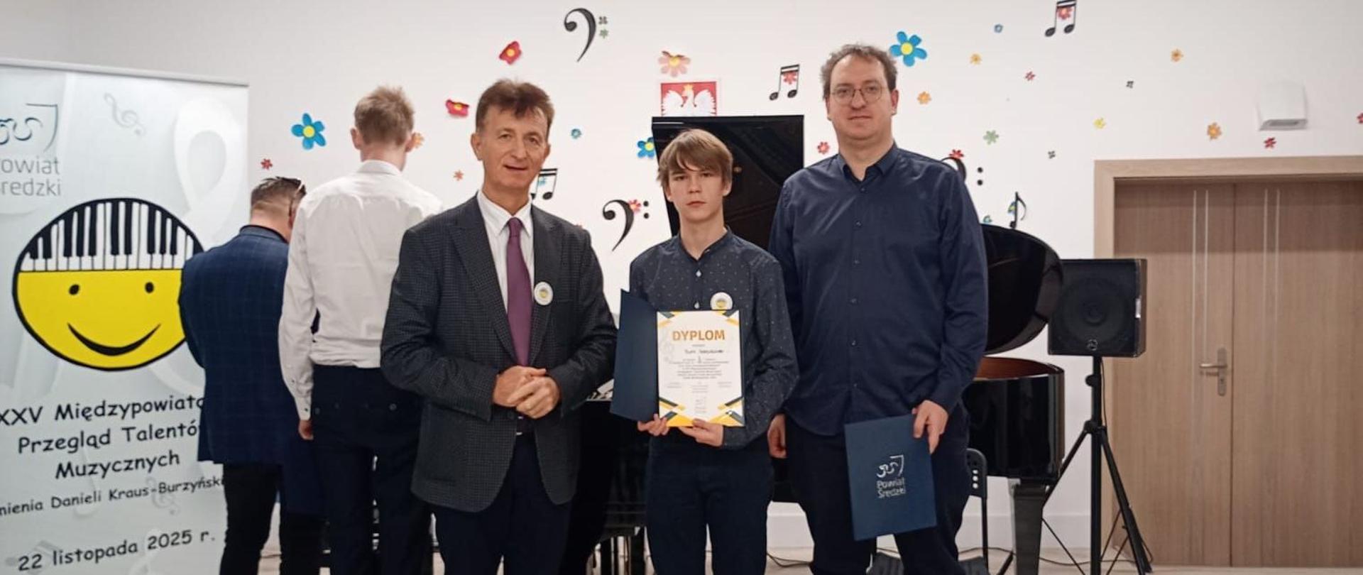 Zdjęcie przedstawia nauczycieli oraz laureata konkursu pozującego z dyplomem za zajęcie drugiego miejsca. W tle banner konkursowy.