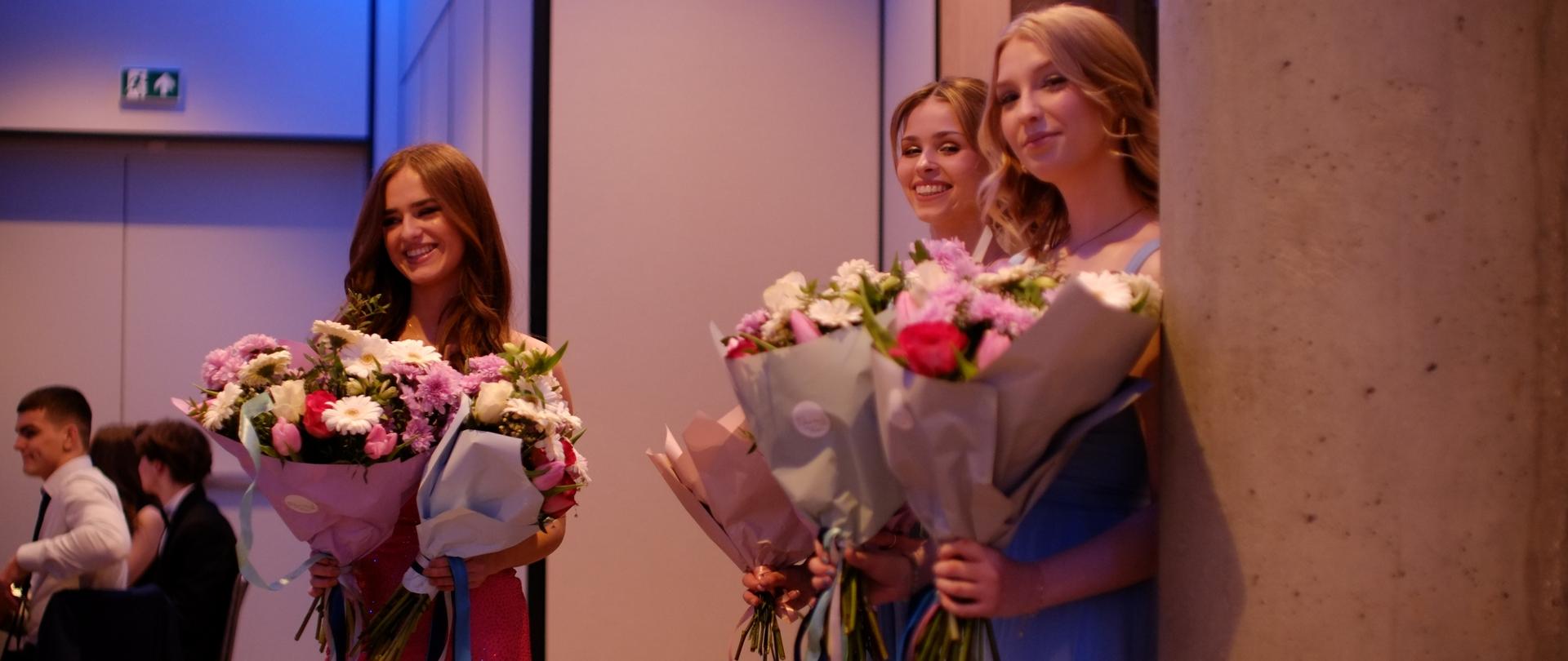 zdjęcie przedstawia uczennice stojące z przygotowywanymi do wręczenia bukietami kwiatów