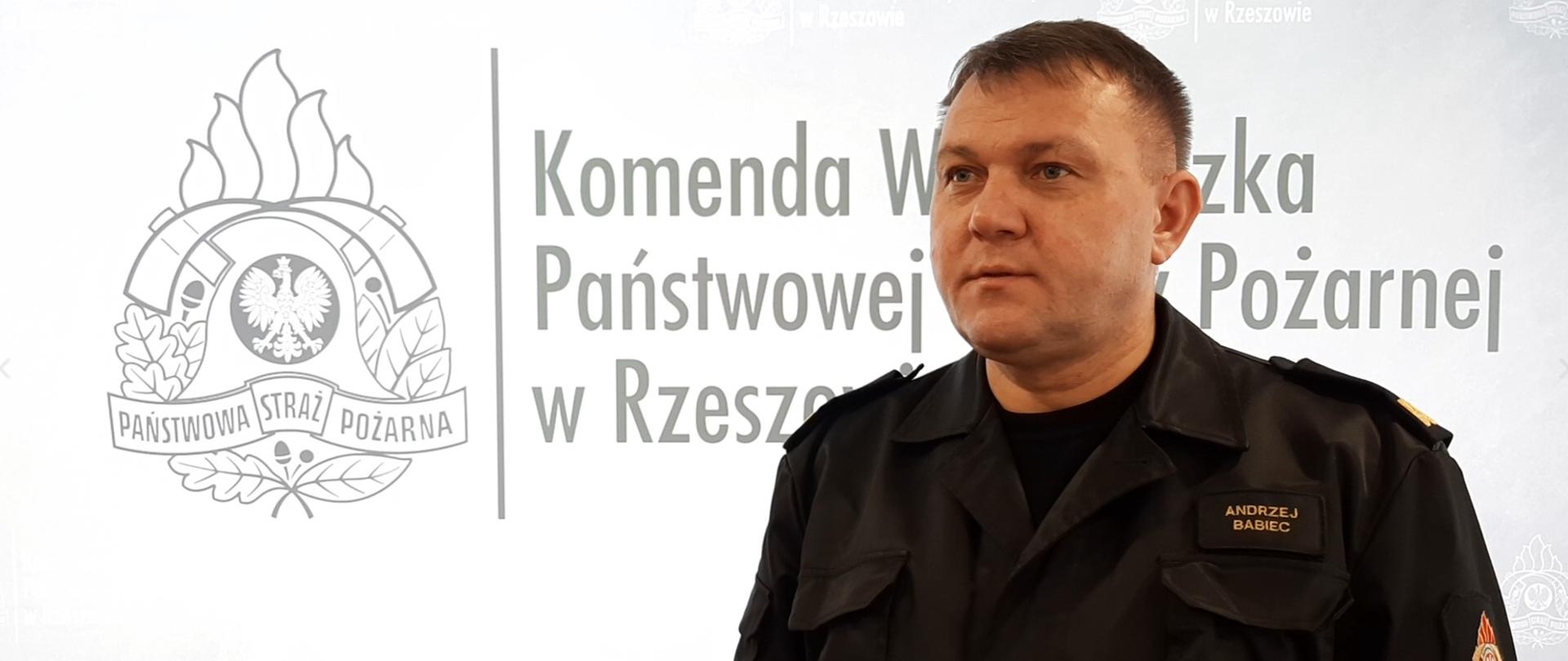 Na zdjęciu podkarpacki komendant wojewódzki Państwowej Straży Pożarnej w mundurze dowódczo-sztabowym. W tle widać szary logotyp Państwowej Straży Pożarnej oraz napis Komenda Wojewódzka Państwowej Straży Pożarnej w Rzeszowie