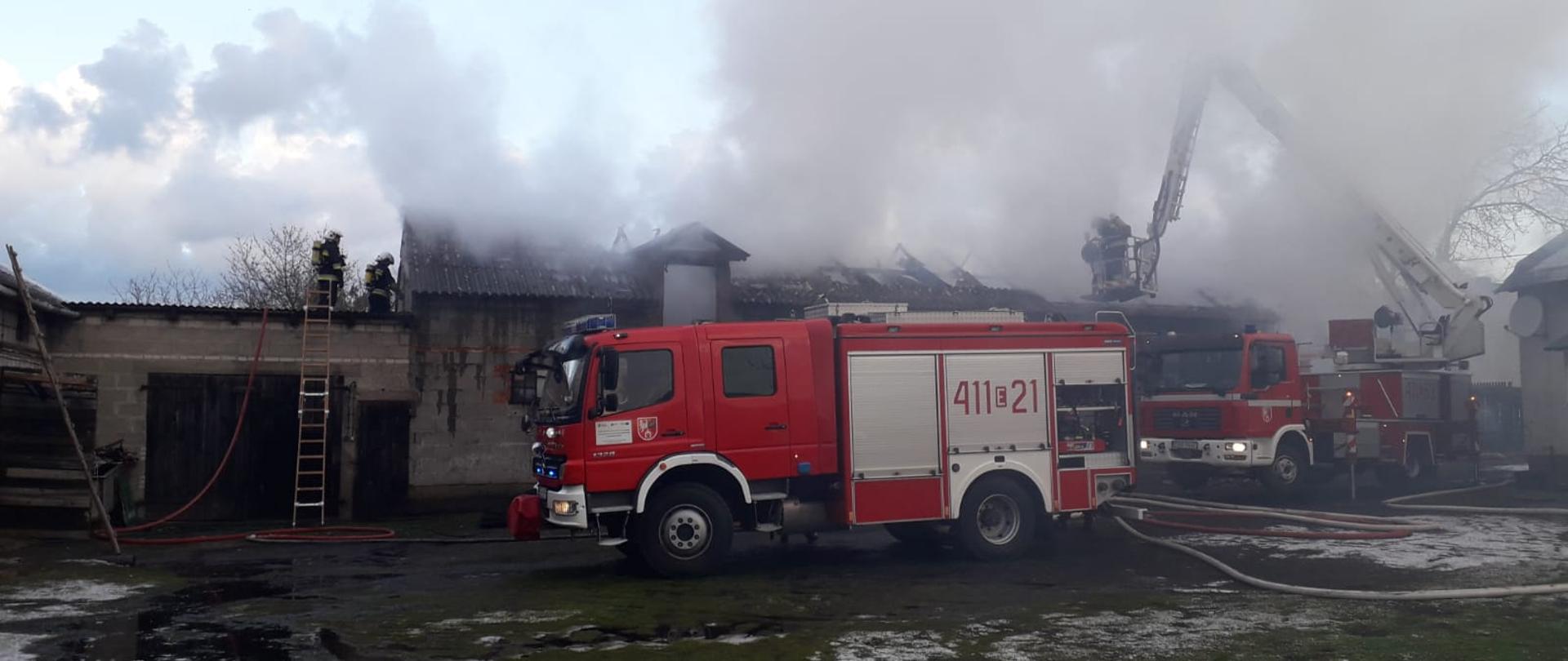 Na zdjęciu rozwinięcie bojowe z udziałem dwóch samochodów pożarniczych; strażacy gaszą palący się budynek prowadząc linie gaśnicze, odpowiednio z poziomu poddasza oraz z podnośnika mechanicznego.