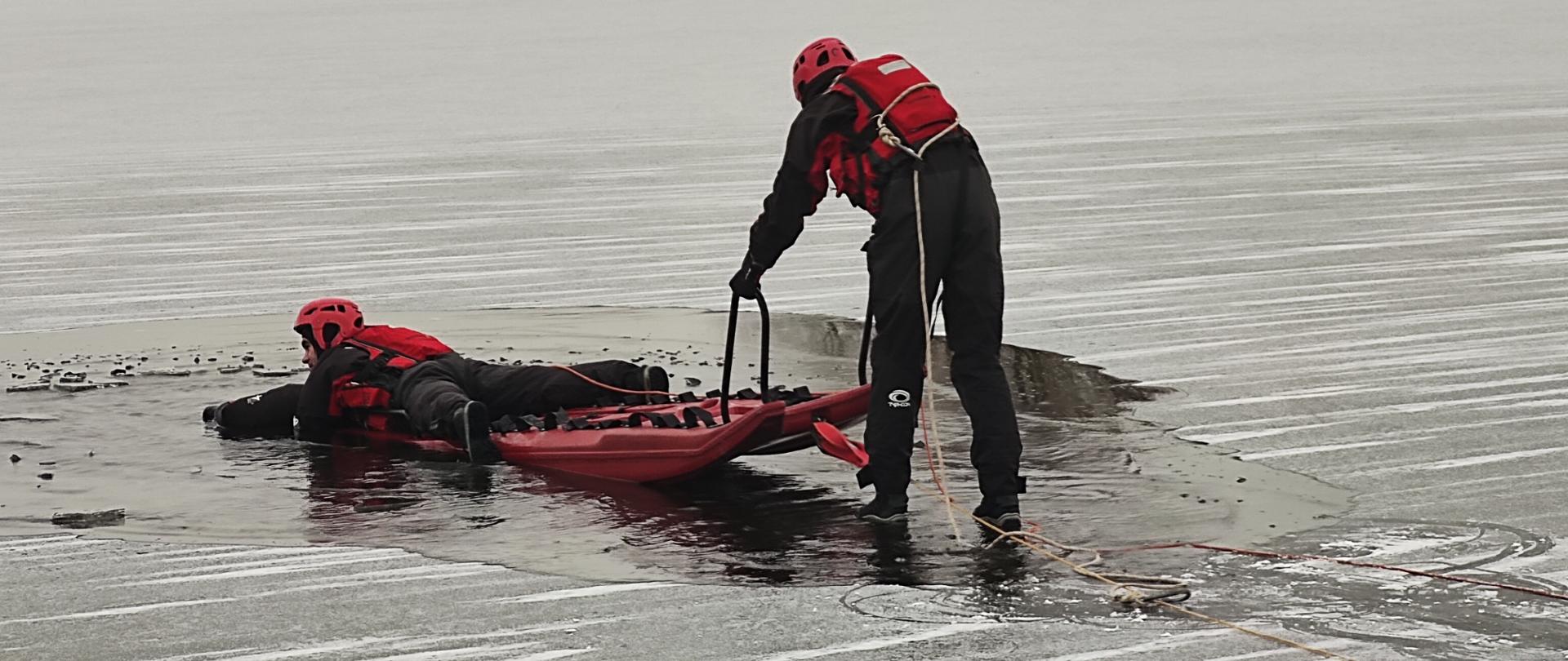 Na zdjęciu dwóch ratowników PSP na saniach lodowych przemieszczających się po cienkim lodzie na akwenie wodnym.
