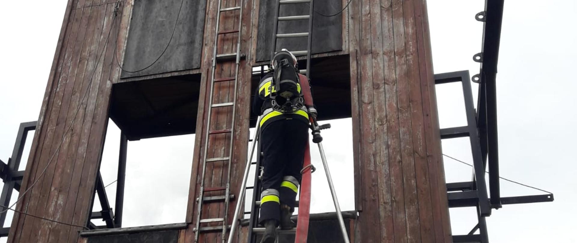 Na zdjęciu widać strażaka na drabinie D10W przed wspinalnią na JRG 1 Kalisz/ Strażak ubrany jest w czarne ubranie specjalne i hełm. Na plecach ma Aparat Ochrony Układu Oddechowego. Drabina ma rozstawione podpory. Po drabinie prowadzona jest linia gaśnicza. W Pobliżu drabiny stoją inni, umundurowani strażacy. Wspinalnia ma kolor brązowo-czarny.