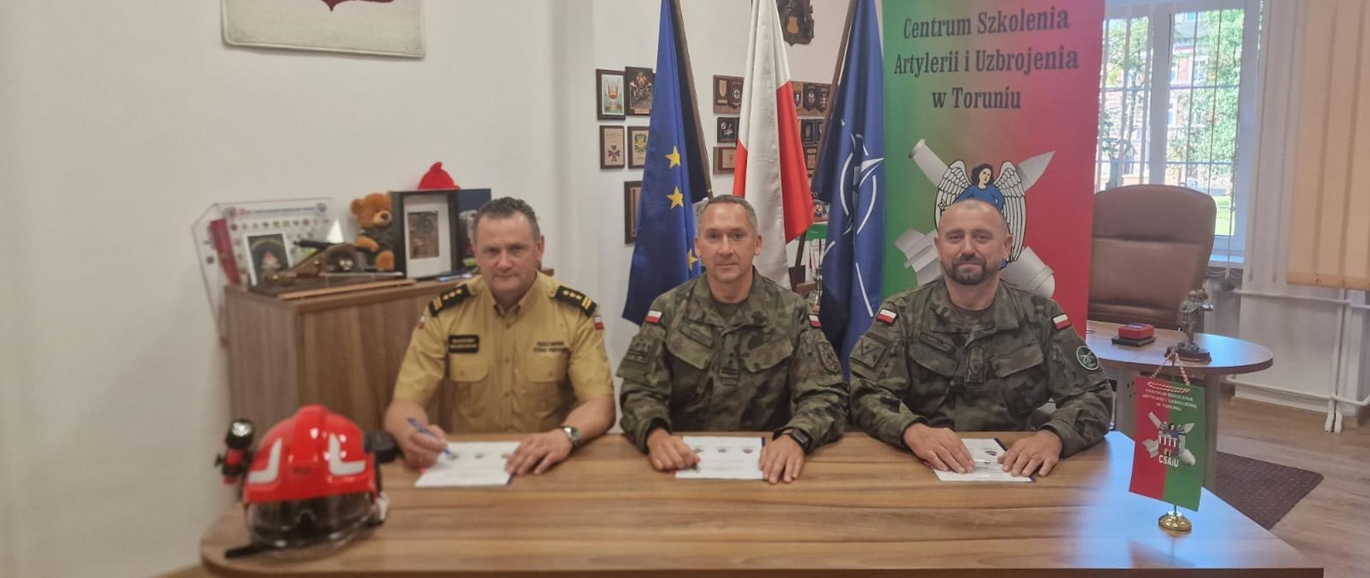 Przy biurku od lewej strony siedzą: Komendant Miejskim Państwowej Straży Pożarnej w Toruniu
st. bryg. Sławomir RESZKOWSKI, Szef Delegatury Wojskowej Ochrony Przeciwpożarowej w Bydgoszczy ppłk Tomasz PAPROCKI, Komendant Centrum Szkolenia Artylerii i Uzbrojenia
płk Remigiusz ZIELIŃSKI, w trakcie podpisywania porozumienia, w tle flagi