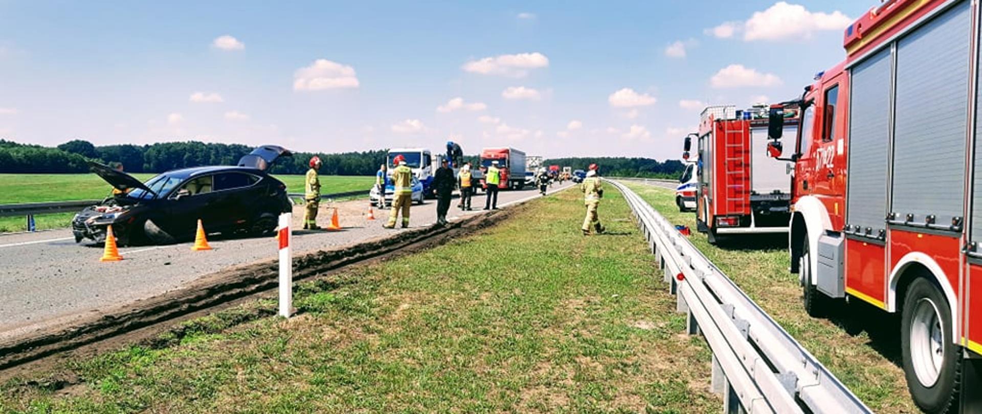 Kolizja na drodze ekspresowej S5, odcinek pomiędzy węzłami Bojanowo-Rawicz. Widok na miejsce kolizji z pasa zieleni oddzielającego jezdnie. Samochód osobowy lexus biorący udział w zdarzeniu. Auto ma podniesioną pokrywę silnika i drzwi bagażnika. Przód samochodu jest uszkodzony. Obok samochodu stoi trzech strażaków oraz rozstawione są pachołki ostrzegawcze. W tle korek samochodów. Z prawej strony bariery energochłonnej karetka pogotowia ratunkowego i dwa wozy straży pożarnej.