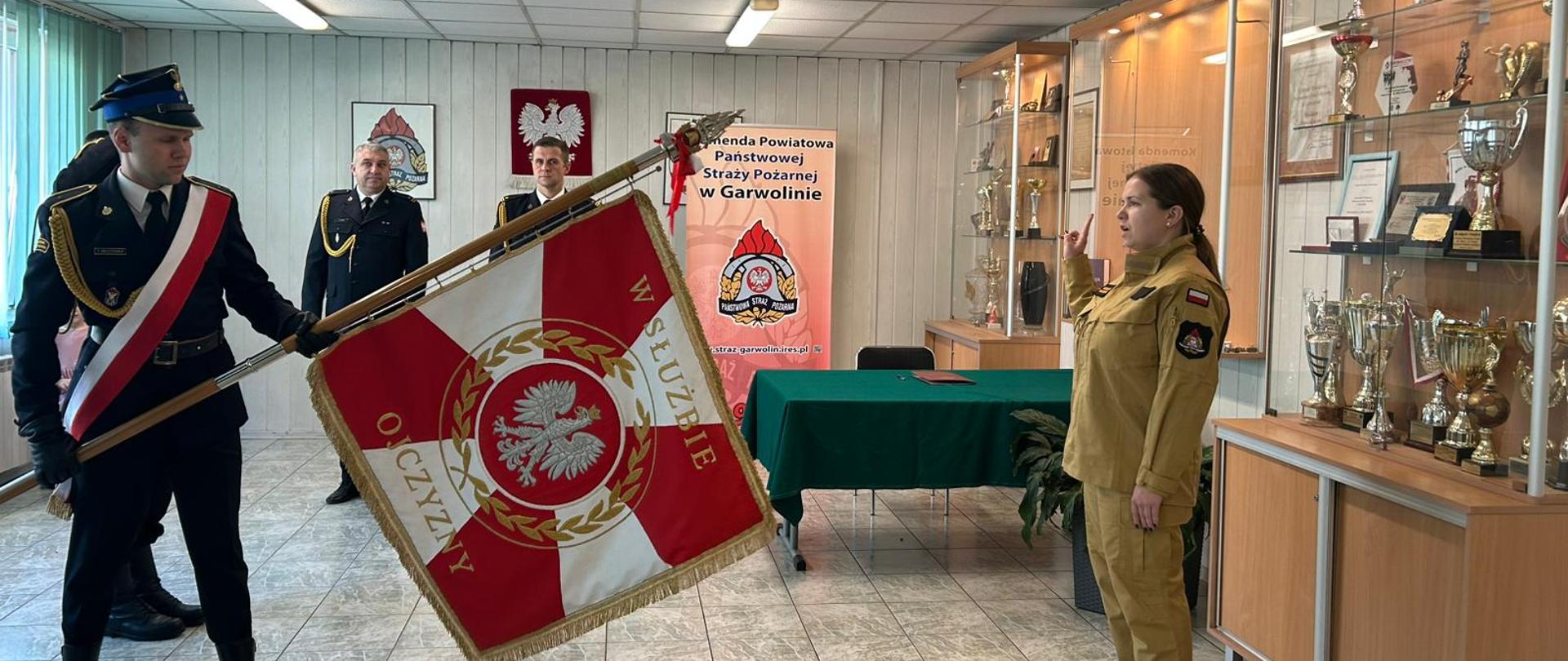 Kobieta w jasnobrązowym mundurze salutuje, stojąc na baczność w pomieszczeniu z licznymi gablotami z trofeami. Przed nią widać oficera w mundurze galowym, który trzyma duży sztandar z orłem i napisem "W SŁUŻBIE OJCZYŹNIE".