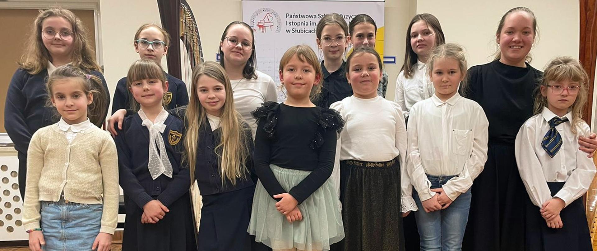 Na zdjęciu stoi grupa dziewczynek i nauczycielka, za nimi stoi harfa.