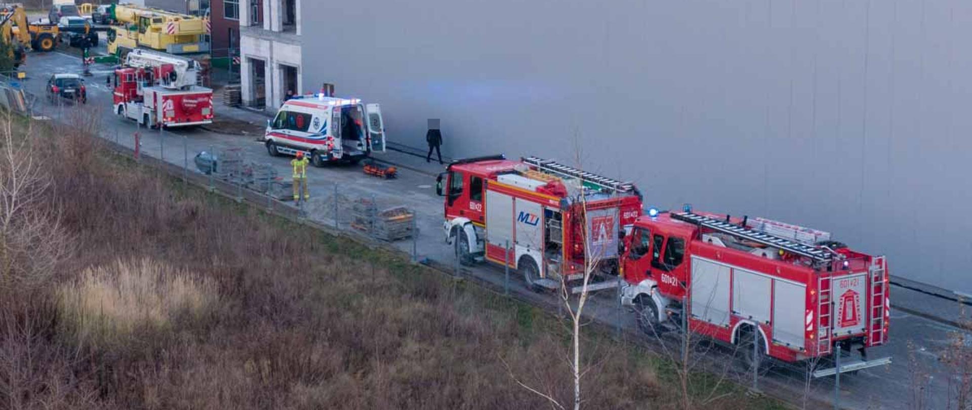 Na zdjęciu 3 pojazdy strażackie w tym podnośnik na niebieskich sygnałach świetlnych, karetka z otwartymi drzwiami stoi przed budynkiem w trakcie budowy
