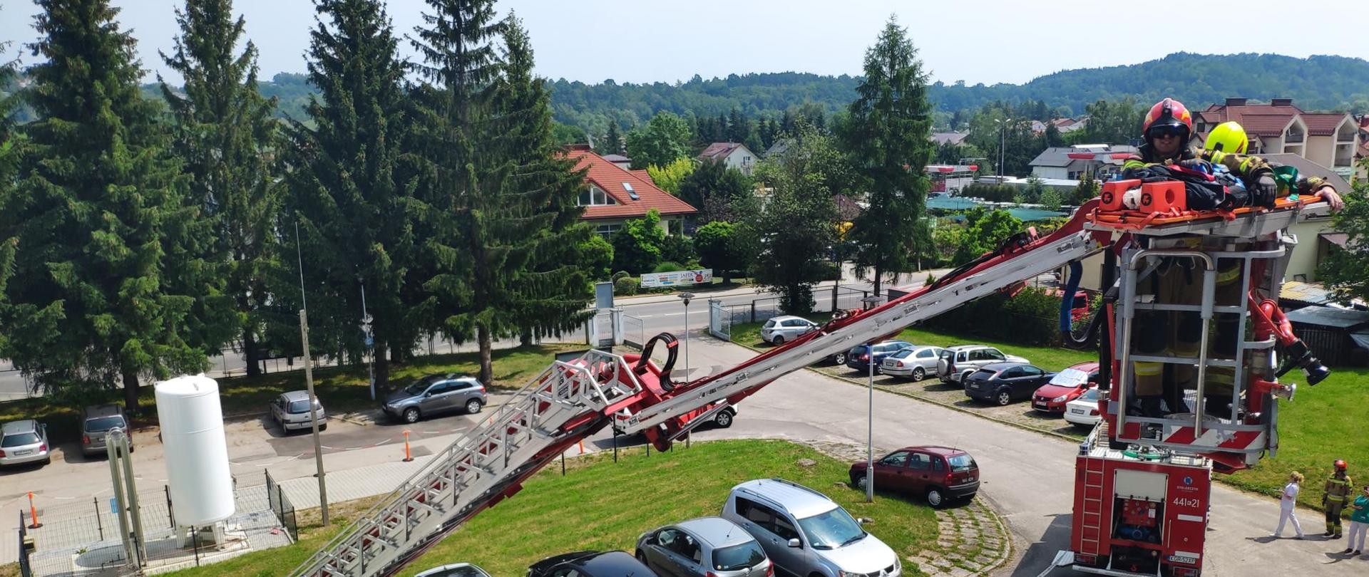 Na zdjęciu widoczni strażacy w ubraniach specjalnych koloru piaskowego jeden w hełmie czerwonym drugi w żółtym w koszu podnośnika hydraulicznego ewakuujący na noszach poszkodowaną osobę podczas ćwiczeń na oddziale geriatrycznym w szpitalu specjalistycznym w Gorlicach.