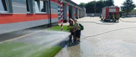 Na zdjęciu strażak, który trzyma wąż obok stoi dziecko ,które leje wodę.