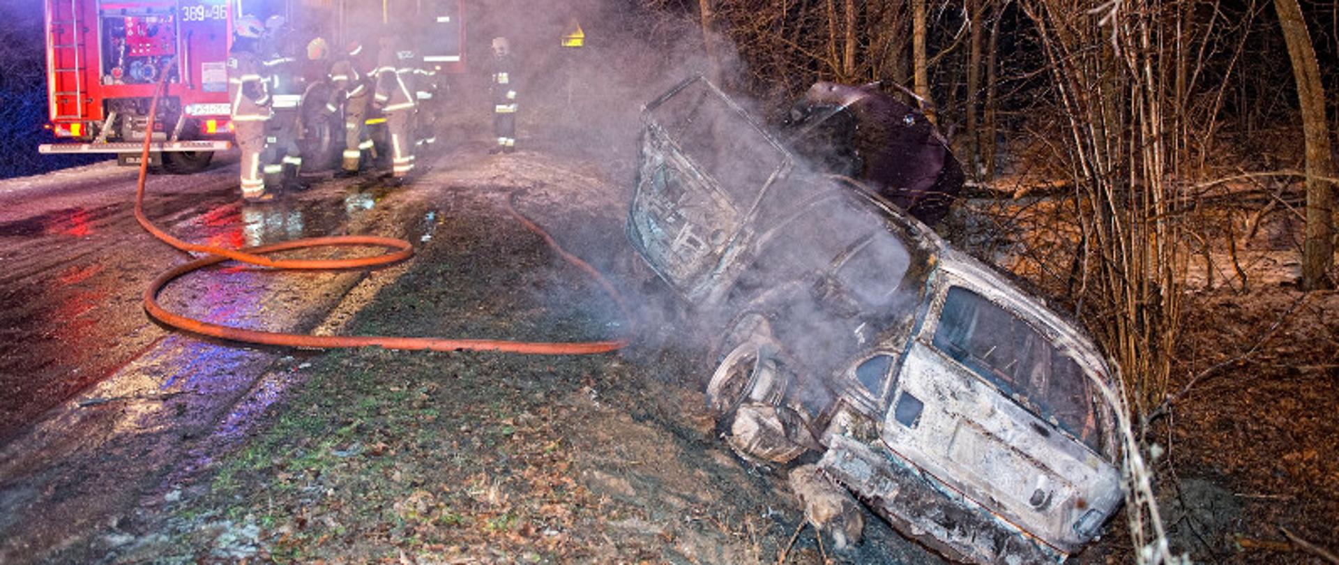 Pożar samochodu osobowego, który znajduje się w rowie obok jezdni. Na zdjęciu spalony wrak pojazdu, w tle strażacy.