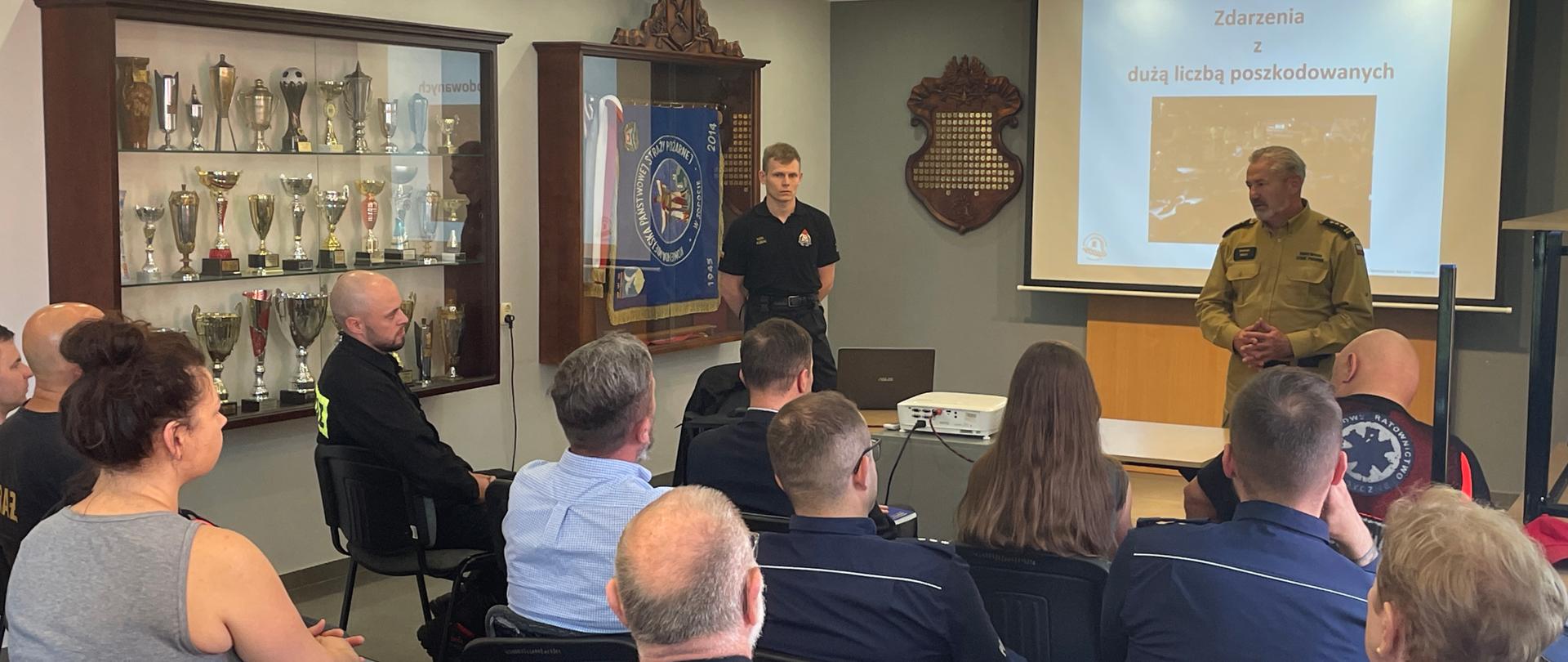 Swietlica komendy. Na ekranie wyświetlona prezentacja. Na krzesłach siedzą uczestnicy spotkania. Przemawia komendant straży