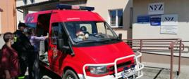 Kolorowe zdjęcie przedstawia na pierwszym planie, czerwony oznakowany samochód lekki Ochotniczej Straży Pożarnej, do którego wsiadają osoby. Po lewej stronie widoczne dwie osoby cywilne. Jedna przy otwartych drzwiach wsiada do samochodu samochodu. Przy niej stoi umundurowany na czarno strażak, asekurujący starszą osobę przy wejściu do samochodu. W tle widoczna część elewacji budynku wielofunkcyjnego, w którym zlokalizowany jest punkt szczepień. Widoczne napisy na budynku NFZ.