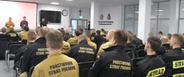 Strażacy w umundurowaniu w kolorze musztardowym oraz czarnym siedzą na krzesłach przed nimi stoją organizator oraz wykładowcy szkolenia.
