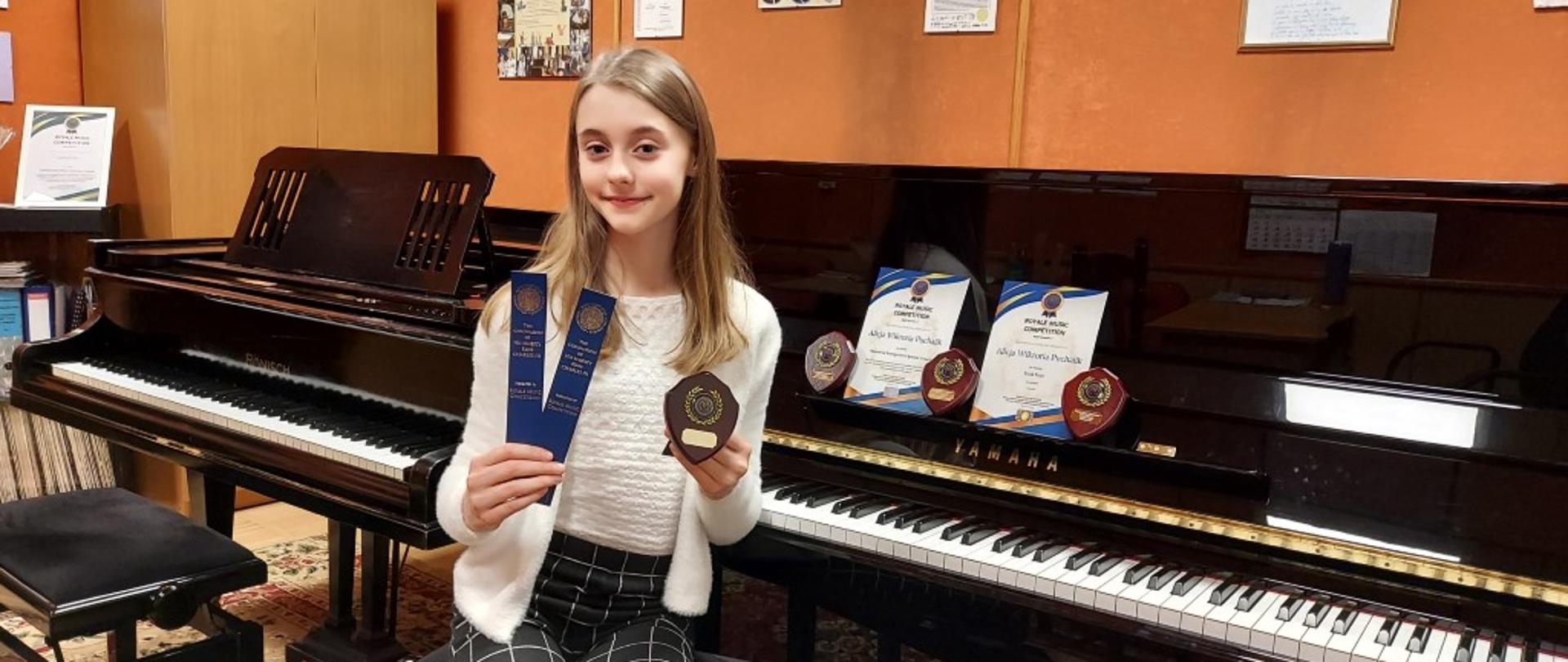 Zdjęcie kolorowe .Dziewczynka o blond włosach w białym sweterku i ciemnych spodniach, siedzi na tle pianina i fortepianu, w jednej ręce trzyma zakładki do książek, a w drugiej trofeum w postaci tarczy. Na pulpicie pianina stoją jeszcze trzy trofea oraz dwa dyplomy. 