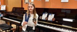 Zdjęcie kolorowe.Dziewczynka o blond włosach w białym sweterku i ciemnych spodniach, siedzi na tle pianina i fortepianu, w jednej ręce trzyma zakładki do książek, a w drugiej trofeum w postaci tarczy. Na pulpicie pianina stoją jeszcze trzy trofea oraz dwa dyplomy. 