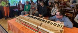 Zdjęcie kolorowe. Grupa uczniów i nauczycieli przygląda się mechanizmowi klawiatury fortepianu. mgr Wojciech Wawryków prezentuje części mechanizmu. W tle wnętrze sali kameralnej PSM w Słupsku.