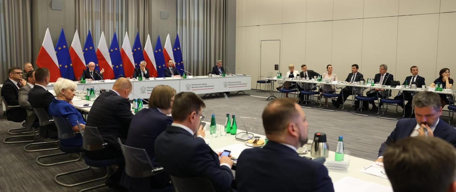 grupa kilkudziesięciu osób siedzi przy okrągłym stole. w tle flagi polskie i unijne