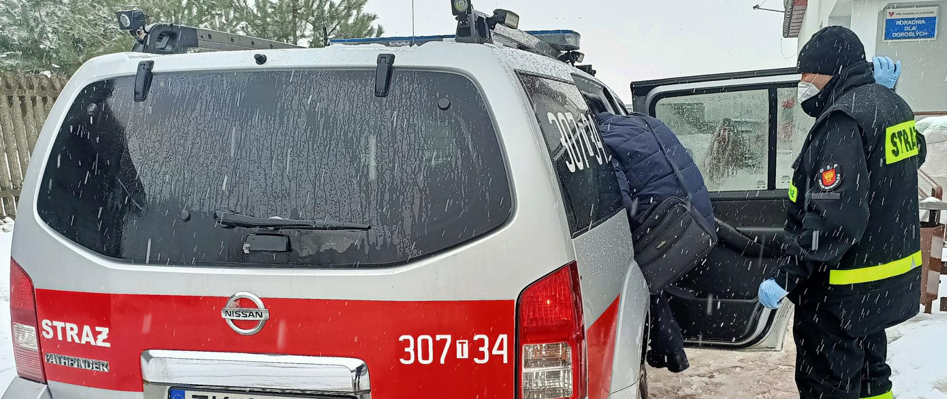 Zdjęcie przedstawia samochód operacyjny Ochotniczej straży Pożarnej z Niewachlowa, do którego wsiada osoba zakwalifikowana do szczepienia. Obok stoi strażak asekurujący starszą osobę.