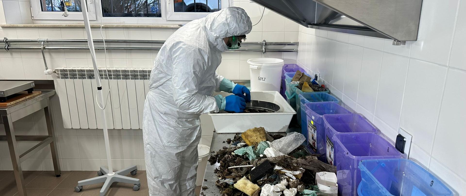 Mężczyzna w białym kombinezonie i masce na twarzy przekłada odpady w białej misce. Obok niego na stole leżą kolorowe pojemniki, a przed nimi sterta odpadów.