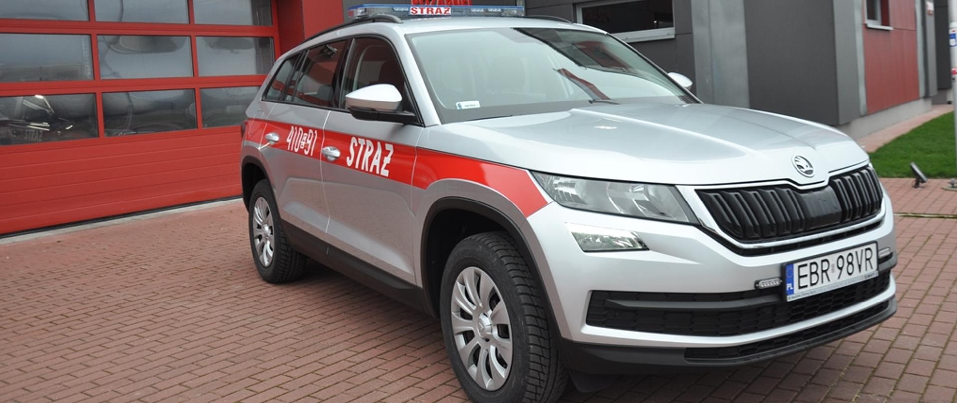 Na zdjęciu samochód lekki operacyjny marki Skoda Kodiaq na tle Komendy powiatowej PSP w Brzezinach. Samochód koloru srebrnego z oznaczeniami STRAŻ oraz numerem operacyjnym 410E91. Przód pojazdu