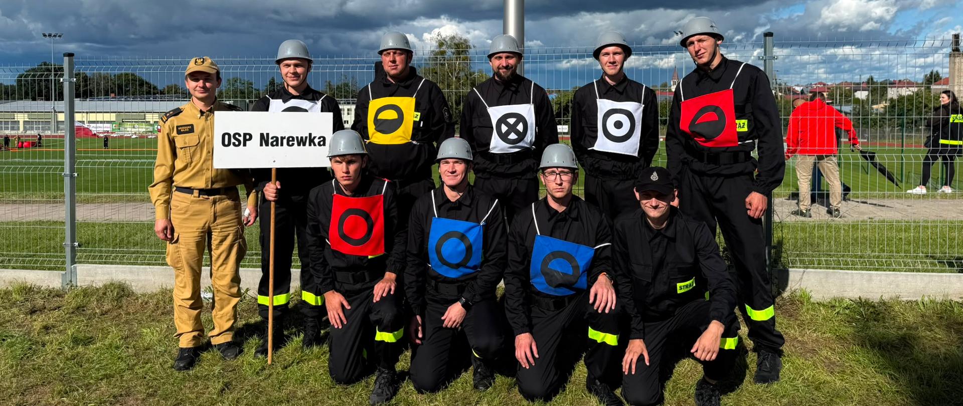 Drużyna strażaków ubrana w ubrania koszarowe i hełmy a także w specjalne oznakowania osób funkcyjnych zgodnie z regulaminem zawodów pozuje do zdjęcia. Czterech strażaków kuca, sześciu stoi zaraz za nimi.. Tło stanowi boisko sportowe.