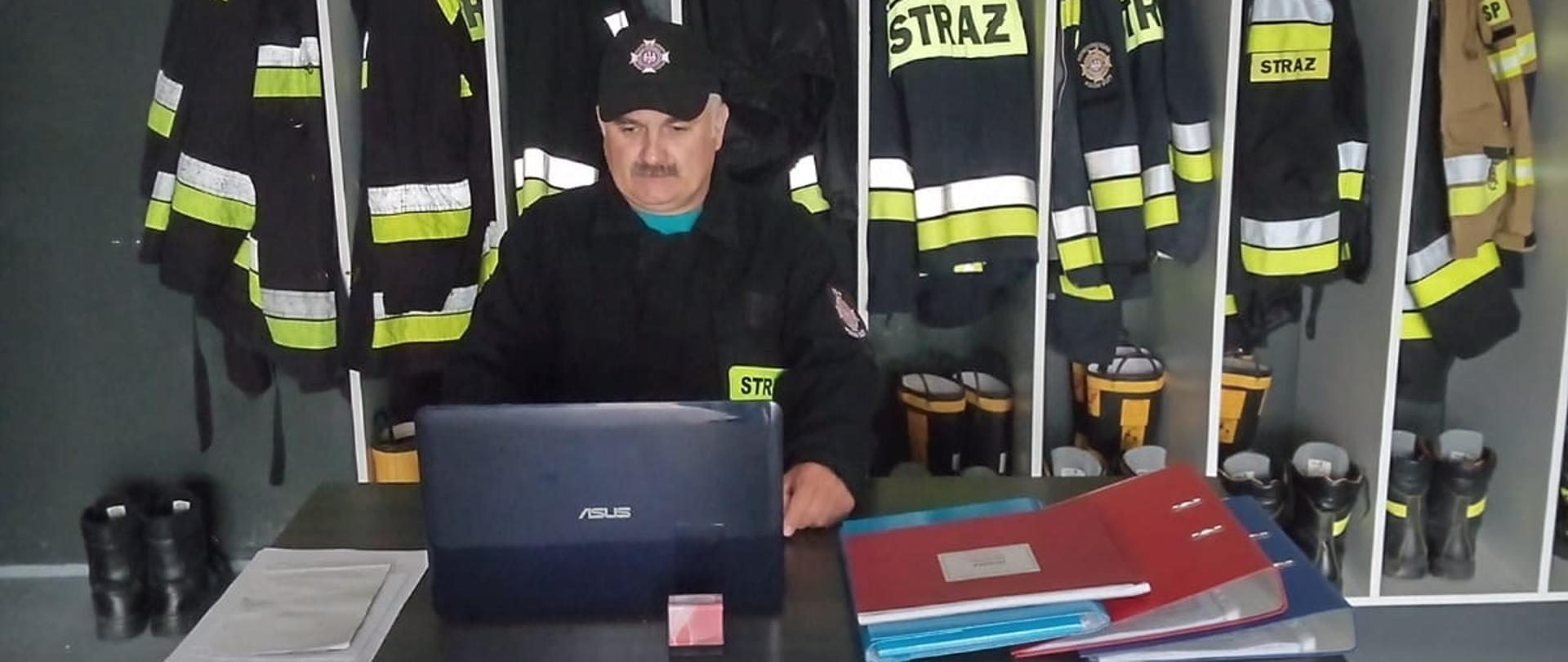 Zdjęcie przedstawia strażaka siedzącego na stołku przed biurkiem ubranego w ubranie koszarowe koloru czarnego, z czapka na głowie koloru czarnego. Strażak ma przed sobą laptop na biurku. Z tyły znajduje się szafka ubraniowa z ubraniami bojowymi oraz hełmami. Na biurku leżą segregatory i dokumenty