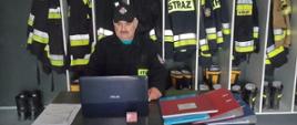 Zdjęcie przedstawia strażaka siedzącego na stołku przed biurkiem ubranego w ubranie koszarowe koloru czarnego, z czapka na głowie koloru czarnego. Strażak ma przed sobą laptop na biurku. Z tyły znajduje się szafka ubraniowa z ubraniami bojowymi oraz hełmami. Na biurku leżą segregatory i dokumenty