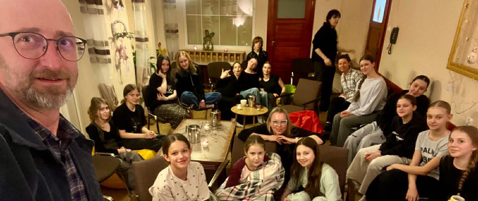 Na pierwszym planie zdjęcia widać mężczyznę w okularach, który patrzy w obiektyw. Na drugim planie uśmiechnięte dziewczyny.