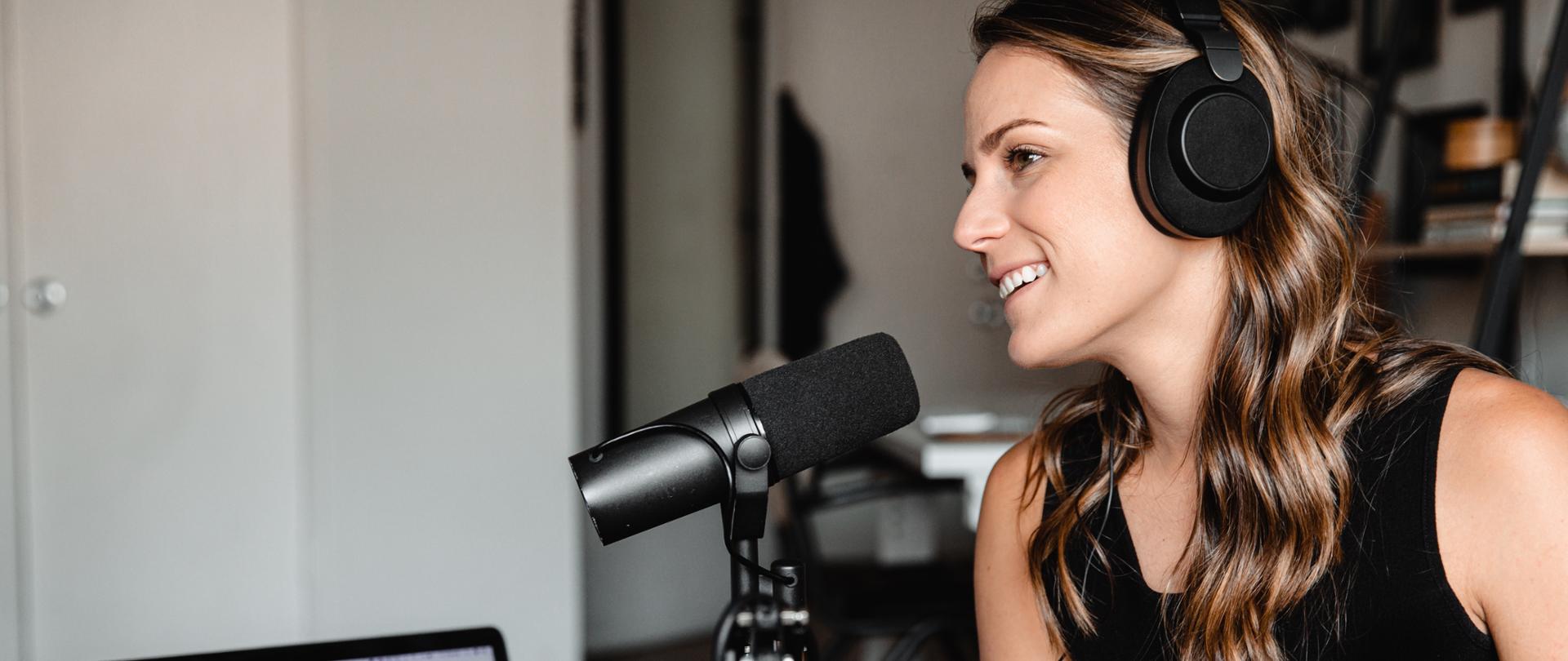 Zdjęcie ilustracyjne w poziomie. ubrana na czarno kobieta siedzi w słuchawkach na uszach. Mówi do stojącego przed nią mikrofonu. Obok na stoliku stoi laptop.