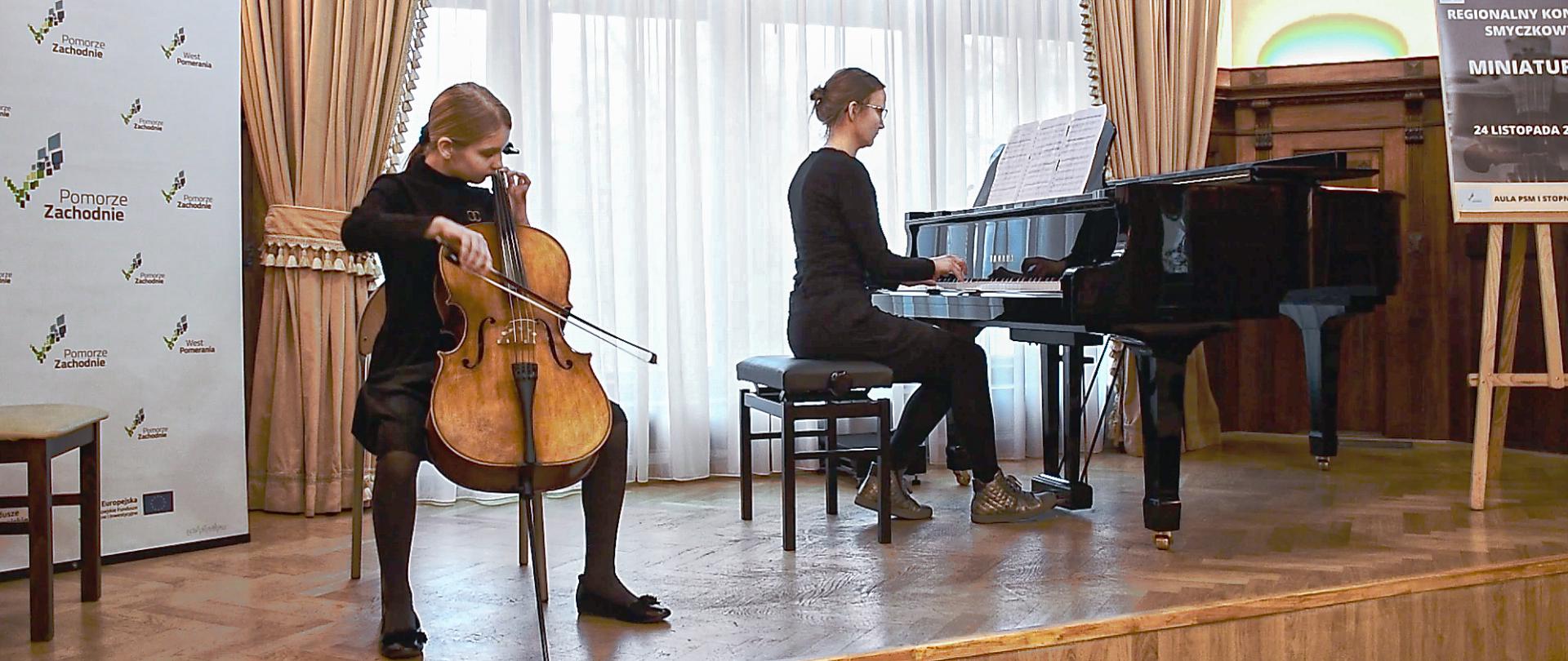 Zdjęcie przedstawia scenę, w tle okno. Na scenie po lewej stronie uczennica grająca na wiolonczeli, po prawej stronie nauczycielka grająca na fortepianie.