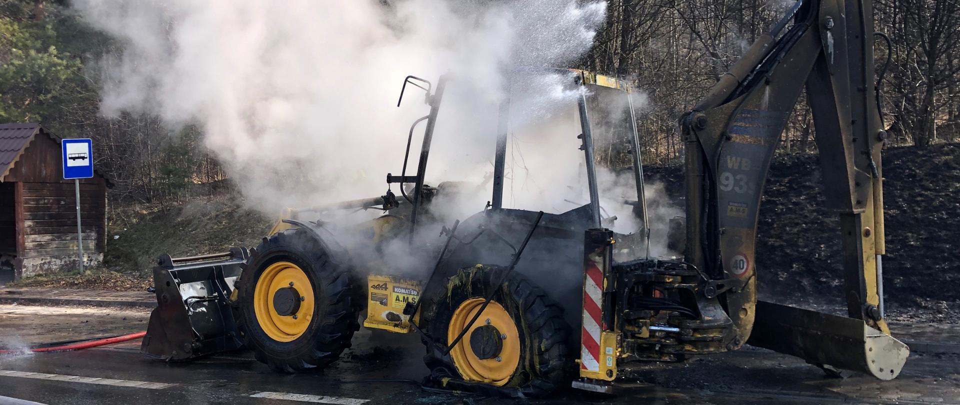 Na zdjęciu przedstawiono spaloną koparkę stojącą na poboczu drogi. Na jezdni leżą nawodnione linie gaśnicze. Wokół urządzenia dym i para powstała z wody podawanej na ognisko pożaru.
