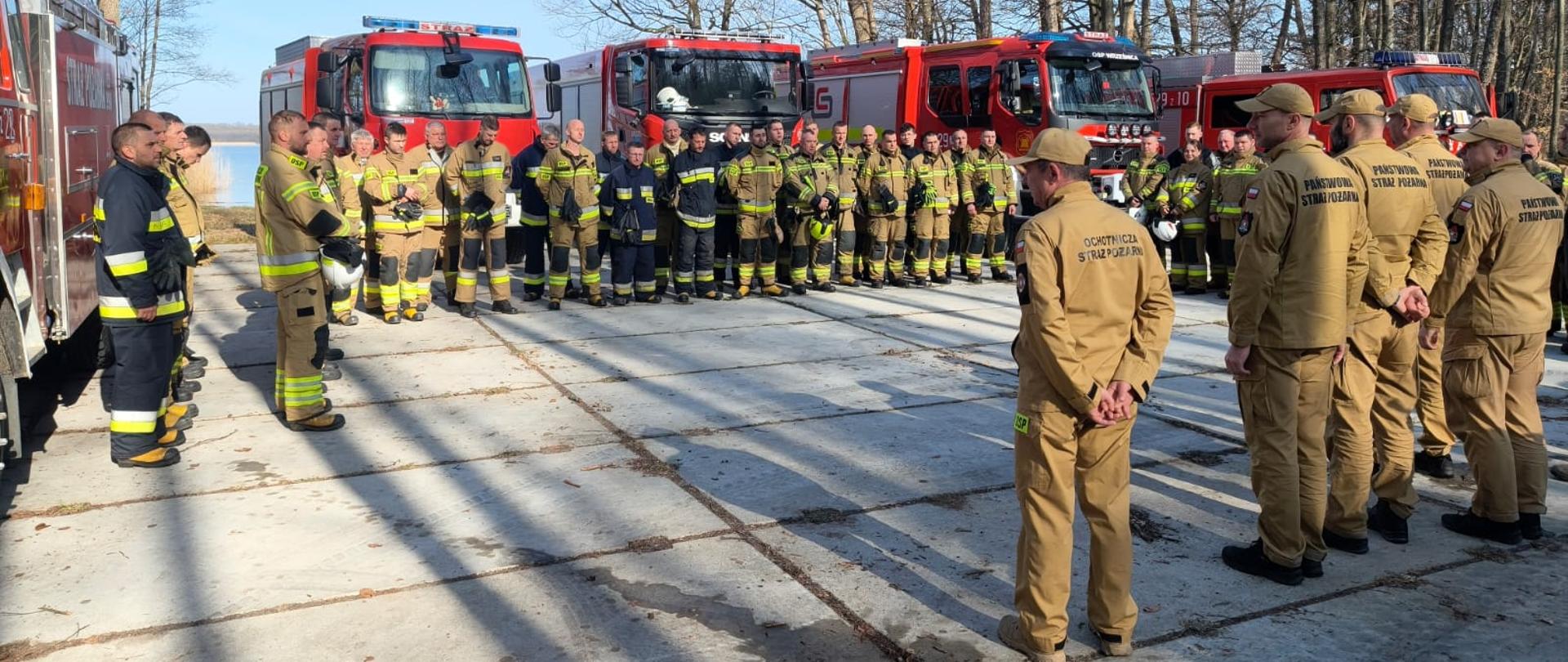 Strażacy ustawieni przed czerwonymi samochodami pożarniczymi podczas odprawy i wspólnego zbiórki na placu przy lesie