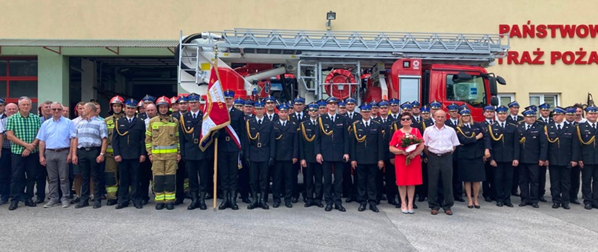 Na placu przed budynkiem strażnicy JRG, na tle drabiny samochodowej do zdjęcia pozują strażacy w umundurowaniu oraz osoby cywilne.
