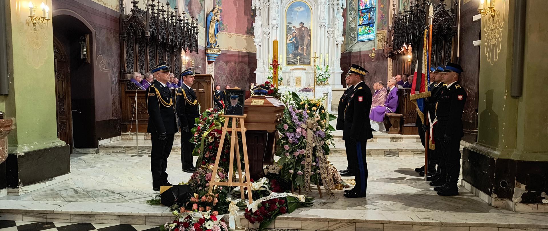 Wnętrze kościoła. Na pierwszym planie trumna z ciałem byłego komendanta PSP w Dębicy. Przy trumnie jest czteroosobowy posterunek wystawiony przez strażaków. Ci strażacy ubrani w mundury galowe stoją na baczność, przy czym dwóch strażaków stoi po prawej stronie i dwóch po lewej stronie trumny. Przy trumnie jest wiele wieńców, kwiatów. Ponadto po prawej stronie, w odległości ok. 2 m od trumny znajduje się sztandar komendy trzymany przez jednego strażaka, ale w asyście dwóch innych. Na drugim planie księżą po jednej i po drugiej stronie ołtarza. W skład liturgicznej służby ołtarza wchodzą także strażacy.