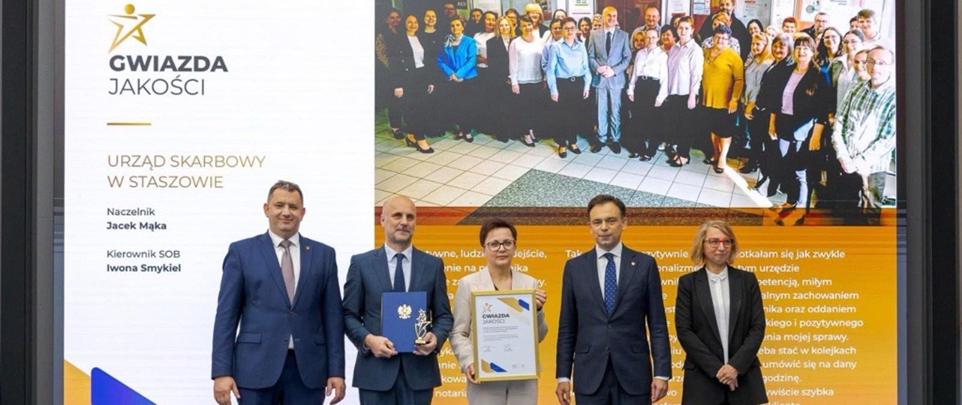 Andrzej Domański, minister finansów i gospodarki, Marcin Łoboda, szef Krajowej Administracji Skarbowej, oraz przedstawiciele Urzędu Skarbowego w Staszowie prezentują dyplom i statuetkę „Gwiazda Jakości”. Na ekranie w tle widoczna jest nazwa urzędu, zdjęcie zespołu oraz fragmenty opinii klientów.