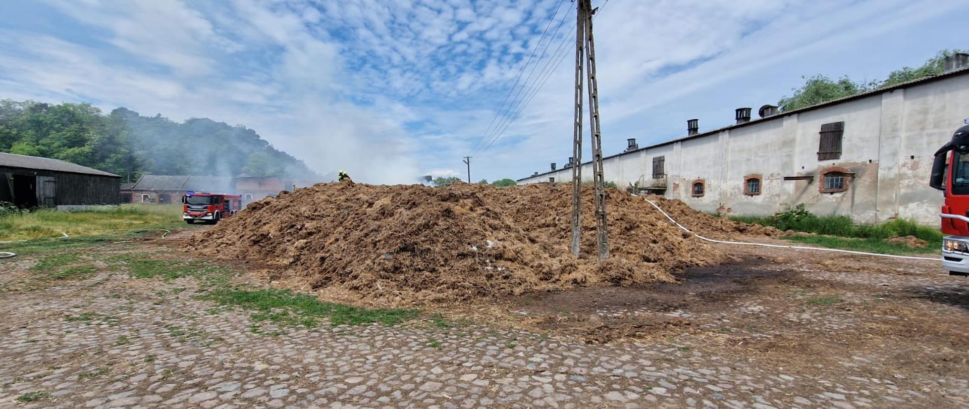 Na środku widać stertę obornika, po lewej samochód strażacki i stodoła, po prawej budynek gospodarczy