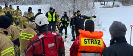 Strażacy stoją obok akwenu wodnego, ubrani są w piaskowe ubrania specjalne oraz niektórzy mają pomarańczowe kamizelki. W tle widać drzewa i śnieg