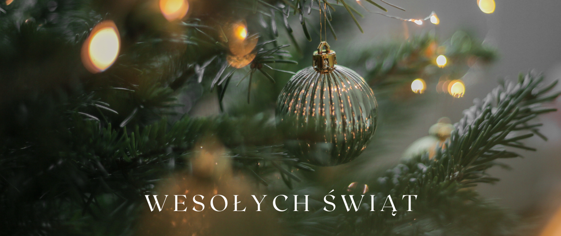 Na obrazku gałązki choinkowe z odbijanymi się światełkami lampek choinkowych. W centralnej części bombka. U dołu obrazka biały napis: "Wesołych Świąt"