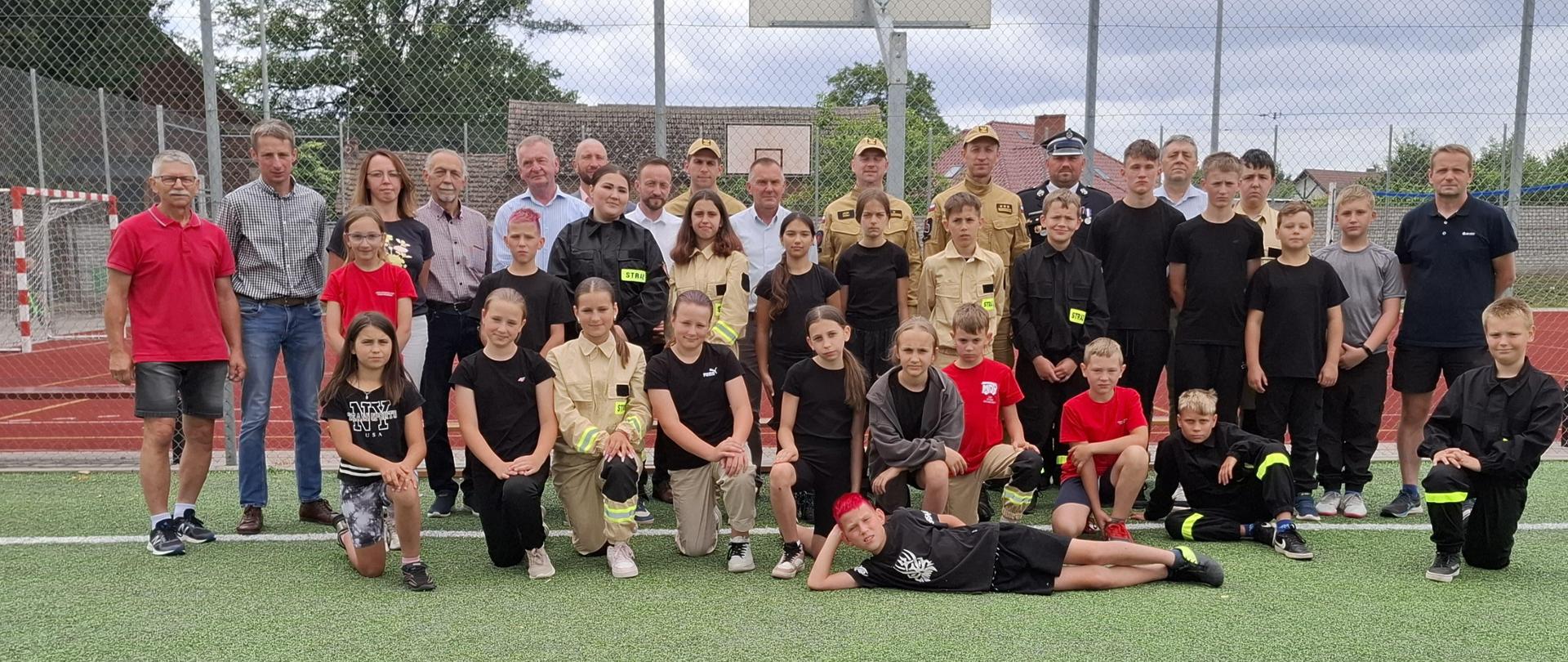 Zdjęcie przedstawia grupę dzieci i młodzieży, uczestników obozu, ustawionej wspólnie z gośćmi na boisku