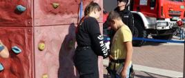 młoda dziewczyna ubrana w czarne spodnie strażackie zakłada szelki ratownicze chłopcu, który będzie wchodził po ściance wspinaczkowej, w tle samochód strażacki oraz strażak ubrany w czarne ubranie koszarowe, widoczne fragmenty budynków Manufaktury