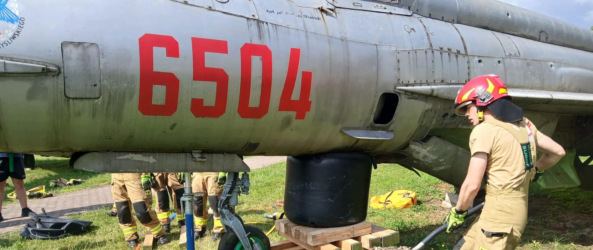 6. Na pierwszym planie widoczna jest część wojskowego samolotu z czerwonym napisem '6504', uniesiona na hydraulicznej podporze i czarnej poduszce niskociśnieniowej. Obok samolotu funkcjonariusz w hełmie i ubraniu ochronnym przenosi ziemię na łopacie. W tle, za samolotem, widoczne są nogi pozostałych uczestników ćwiczeń