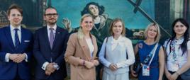 Sześć osób stoi blisko siebie i pozuje do zdjęcia na tle dużego, kolorowego muralu. Po lewej stronie stoi młody mężczyzna w okularach, ciemnoniebieskim garniturze i niebieskim krawacie w białe kropki. Obok niego stoi mężczyzna z brodą i okularach, w ciemnym garniturze i wzorzystym krawacie.
Trzecia od lewej stoi kobieta z blond włosami spiętymi do tyłu, ubrana w beżowy garnitur i białą bluzkę. Obok niej stoi kobieta z jasnymi włosami, w jasnym, eleganckim komplecie z wiązaną bluzką. Piąta od lewej stoi kobieta z blond włosami, w niebieskim kombinezonie i z akredytacją na szyi. Po prawej stronie stoi młoda kobieta z długimi, ciemnymi włosami, ubrana w białą bluzkę z wzorzystą chustą na szyi i ciemną spódnicę, z torebką przewieszoną przez ramię.
Mural za nimi przedstawia postać historyczną, Mikołaja Kopernika, z elementami astronomicznymi i architektonicznymi w tle. 