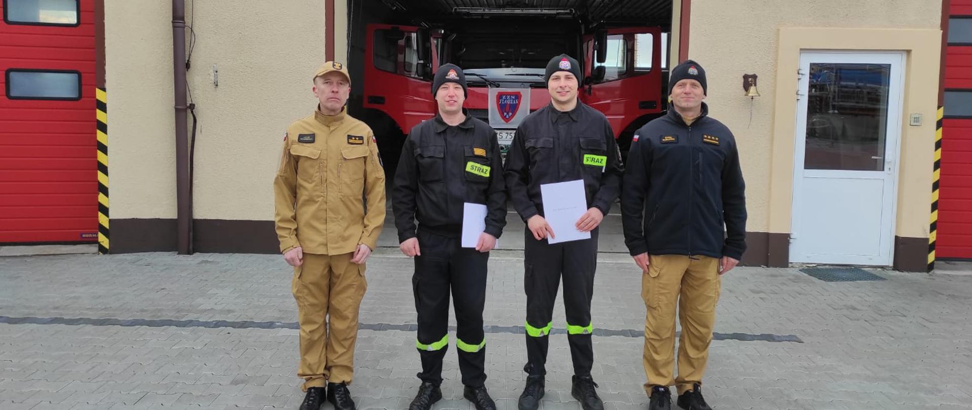 Zdjęcie przedstawia czterech strażaków przed bramą garażową pozujących do zdjęcia.
